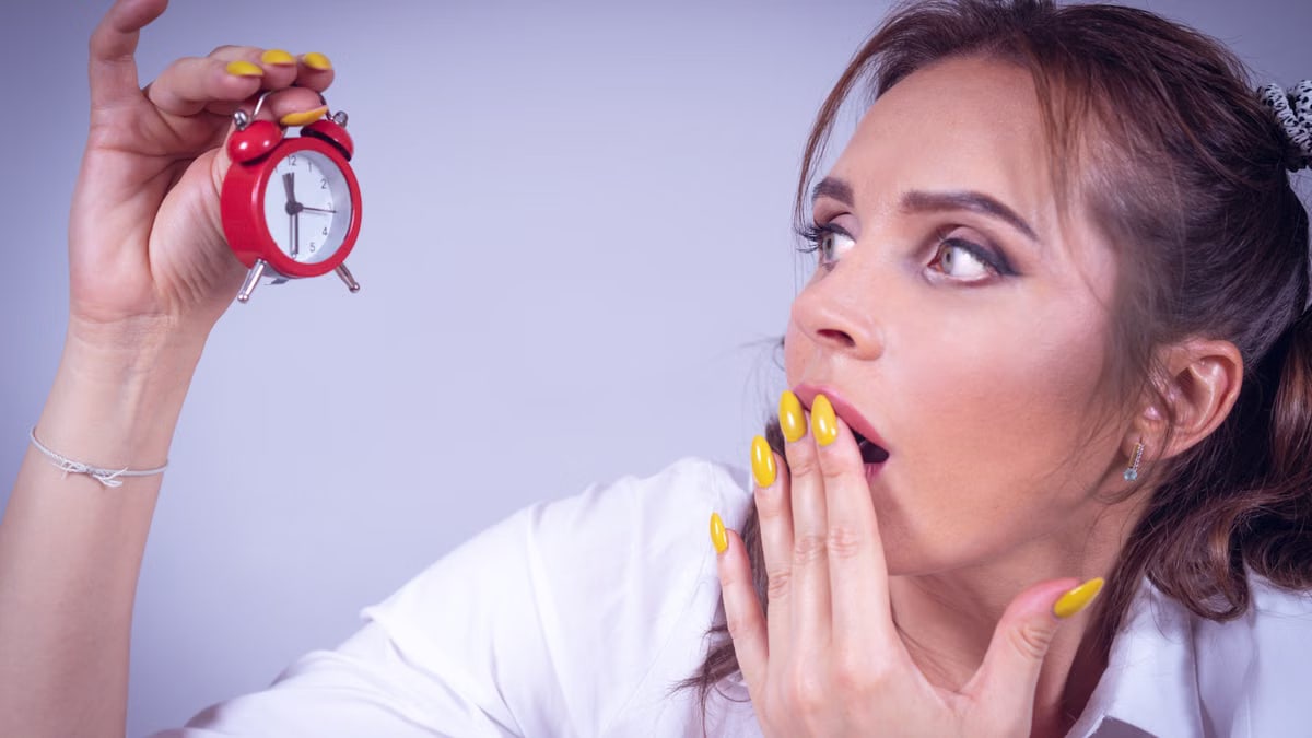 A surprised woman with yellow nails holds a small red alarm clock against a white backdrop.
